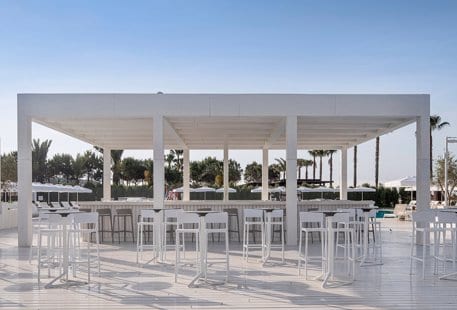 Adult Pool Bar white wooden decking, sleek high-top tables, and a slatted roof structure under a clear blue sky at Voyage Belek Golf & Spa by Kenwood Travel