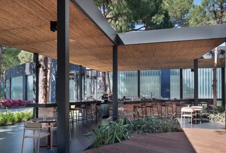 Coffee House natural reed-textured ceiling, sleek black structural columns, and a stone-clad bar counter surrounded by lush pine trees and contemporary seating at Voyage Belek Golf & Spa by Kenwood Travel
