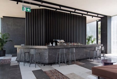 Lobby Bar grey marble counter, sleek grey bar stools, and a black slatted accent wall under industrial track lighting at Voyage Belek Golf & Spa by Kenwood Travel