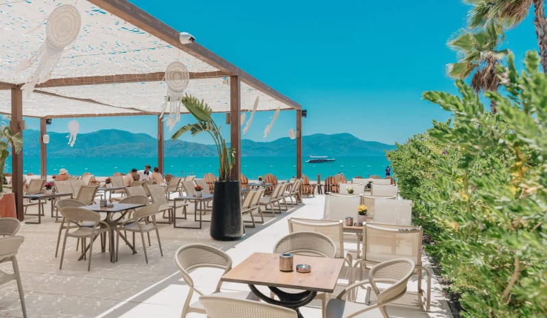 Patara Beach Bar  white modern furniture, a wooden pergola with dreamcatchers, and a panoramic view of the turquoise Mediterranean sea and mountains at Liberty Fabay by Kenwood Travel