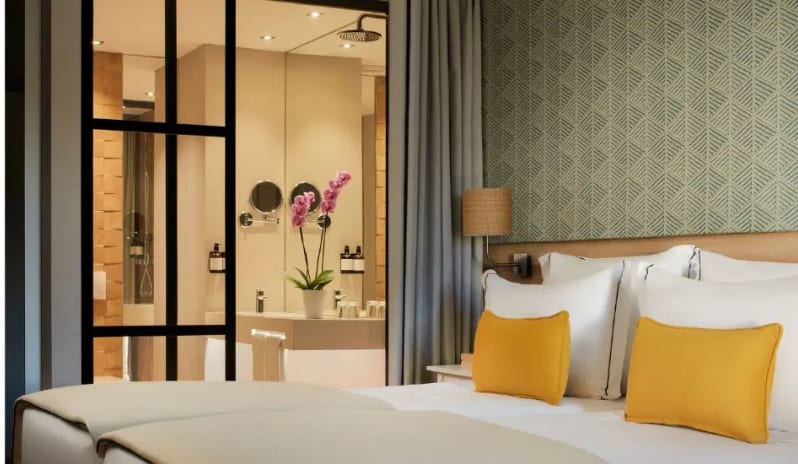 Deluxe Room interior bed with yellow pillows and a textured green accent wall, looking through to a modern ensuite bathroom with a large vanity mirror at Tivoli Alvor Algarve by Kenwood Travel
