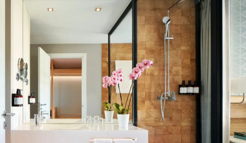 Deluxe Room bathroom interior with a white vanity, chrome fixtures, and a glass-enclosed shower area showcasing warm wood-textured wall tiles at Tivoli Alvor Algarve by Kenwood Travel