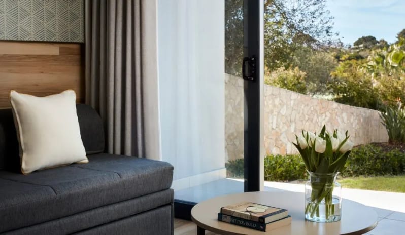 Deluxe Room sitting area in a hotel guest room featuring a round coffee table with white tulips and books, overlooking a stone-walled garden at Tivoli Alvor Algarve by Kenwood Travel