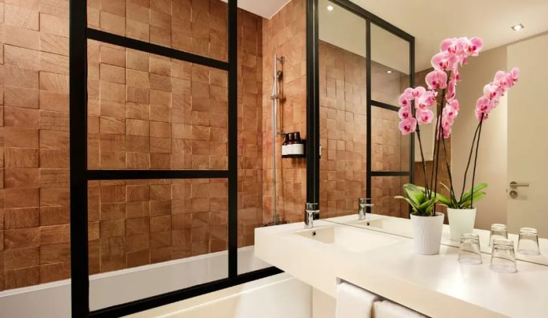 Family Deluxe Room a wood-textured tiled shower wall, black-framed glass partition, white vanity with pink orchids, and large mirrors at Tivoli Alvor Algarve by Kenwood Travel