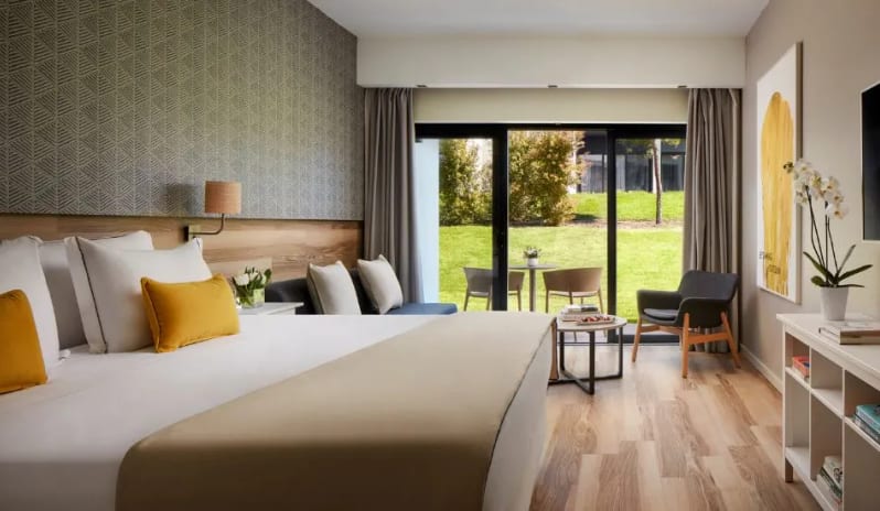 Premium Two-Bedroom large bed with yellow accent pillows, a seating area, and sliding glass doors opening to a green garden patio at Tivoli Alvor Algarve by Kenwood Travel