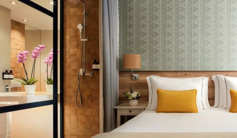 Premium Two-Bedroom bed with yellow pillows next to a glass-walled bathroom with wood-textured tiles and a modern walk-in shower at Tivoli Alvor Algarve by Kenwood Travel