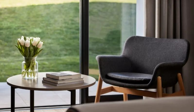 Premium Two-Bedroom dark grey upholstered armchair with wooden legs sitting next to a round side table with white tulips and books, positioned in front of a floor-to-ceiling window overlooking a lawn at Tivoli Alvor Algarve by Kenwood Travel