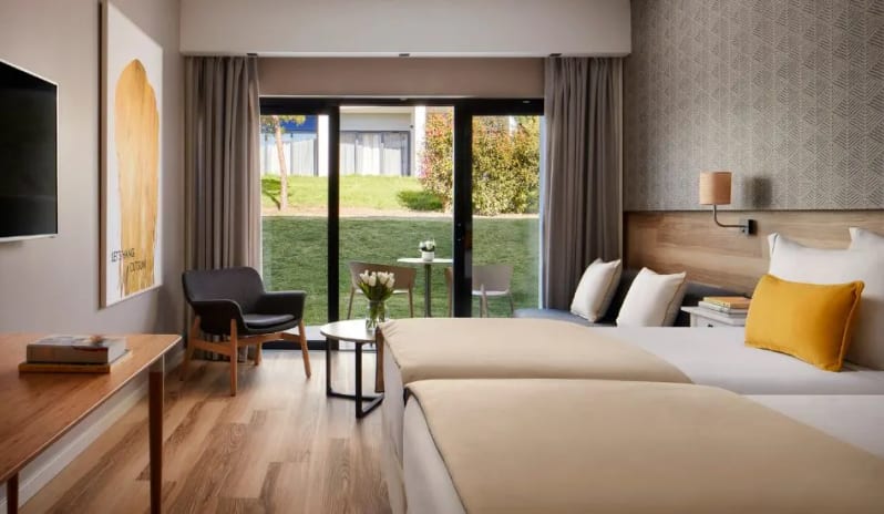 Premium Two-Bedroom large bed with yellow accent pillows, a seating area, and sliding glass doors opening to a green garden patio at Tivoli Alvor Algarve by Kenwood Travel
