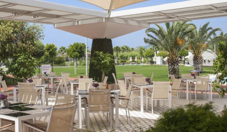 Breeze Snack Restaurant & Bar white tables and chairs under a large tan sail canopy, surrounded by palm trees and manicured green lawns at Hilton Dalaman Sarigerme Resort & Golf by Kenwood Travel