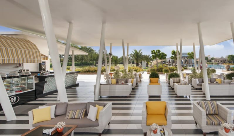 Citrus Coffee & Bar black and white striped flooring, white wicker sofas with yellow cushions, and a sleek shaded canopy at Hilton Dalaman Sarigerme Resort & Golf by Kenwood Travel
