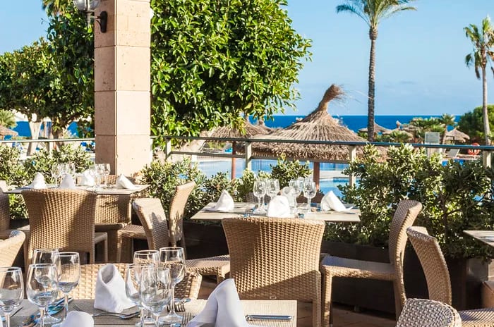 Aire Restaurant – Mediterranean Cuisine woven chairs, set tables with white linens, and a view of the Mediterranean Sea and palm trees at Insotel Punta Prima Resort & Spa by Kenwood Travel