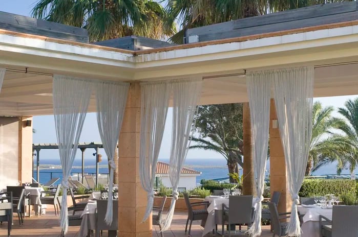 Bar Salón Magón Elegant outdoor Mediterranean restaurant terrace with white string curtains, palm trees, and a scenic ocean view at Insotel Punta Prima Resort & Spa by Kenwood Travel