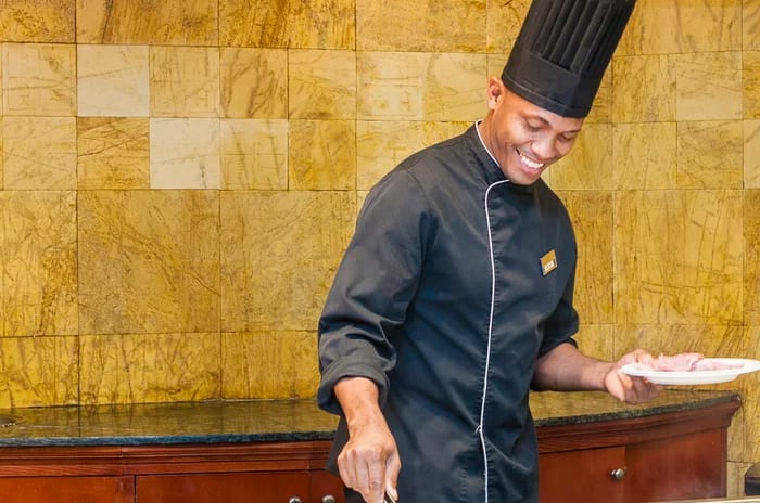 Grill Xoroi Restaurant Smiling chef in a black uniform and tall hat grilling thin slices of meat on a flat-top teppanyaki grill in a restaurant kitchen at Insotel Punta Prima Resort & Spa by Kenwood Travel