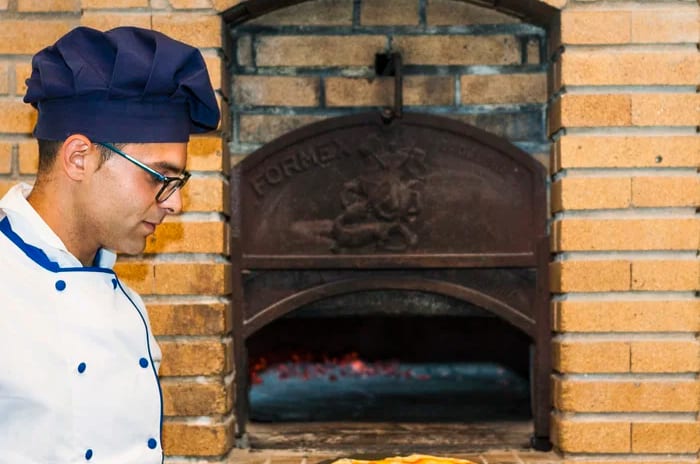 Italian Restaurant Il Faro A chef in a white uniform and blue hat using a pizza peel to remove a fresh thin-crust pizza from a traditional brick wood-fired oven at Insotel Punta Prima Resort & Spa by Kenwood Travel