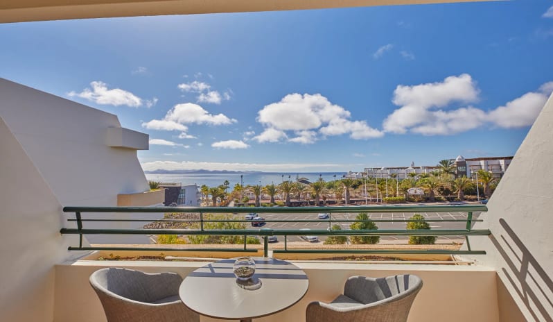 Double Side Ocean View terrace with a small table and two grey chairs overlooking a sunny coastline with palm trees and a blue ocean at Dreams Lanzarote Playa Dorada Resort & Spa by Kenwood Travel