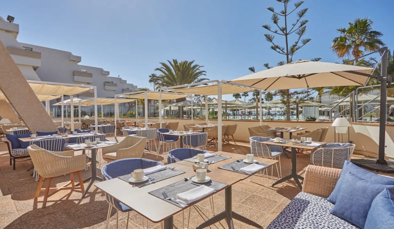 World Cafe rmodern dining tables, blue cushions, and white sun canopies set against a backdrop of palm trees and clear blue sky at Dreams Lanzarote Playa Dorada Resort & Spa by Kenwood Travel