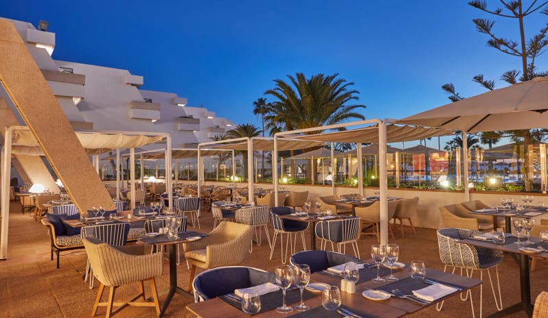 World Cafe outdoor restaurant terrace at twilight featuring modern dining tables with blue seating under white pergolas and palm trees at Dreams Lanzarote Playa Dorada Resort & Spa by Kenwood Travel