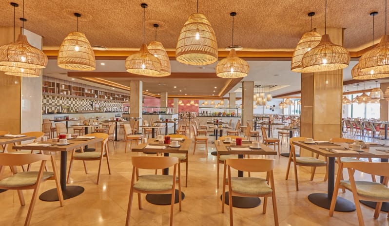 World Cafe restaurant interior featuring wooden dining tables, cushioned chairs, and multiple woven wicker pendant lights over a polished marble floor at Dreams Lanzarote Playa Dorada Resort & Spa by Kenwood Travel