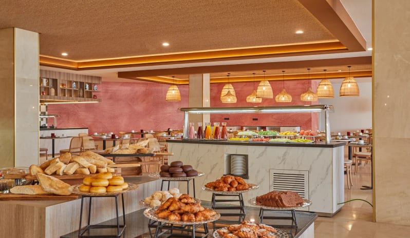 World Cafe breakfast buffet station featuring a variety of fresh pastries, breads, and juices displayed on marble counters in a modern dining room at Dreams Lanzarote Playa Dorada Resort & Spa by Kenwood Travel