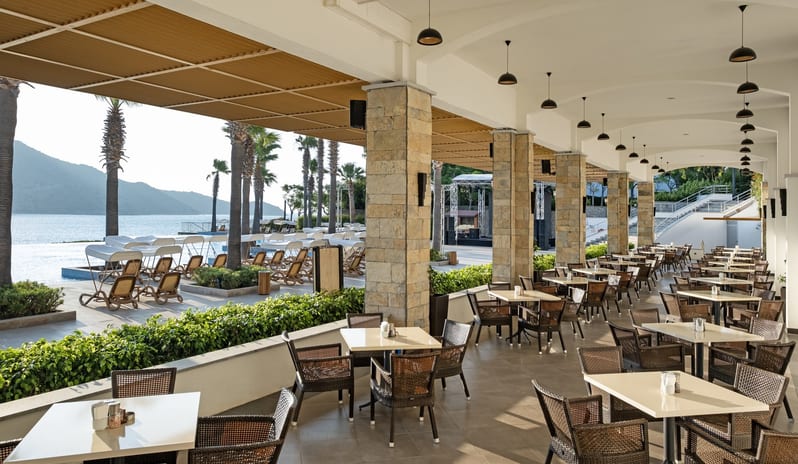 Xanadu Bar An elegant outdoor dining terrace at a seaside resort featuring stone pillars, wicker seating, and a view of a pool and mountains by the ocean at Xanadu Island by Kenwood Travel