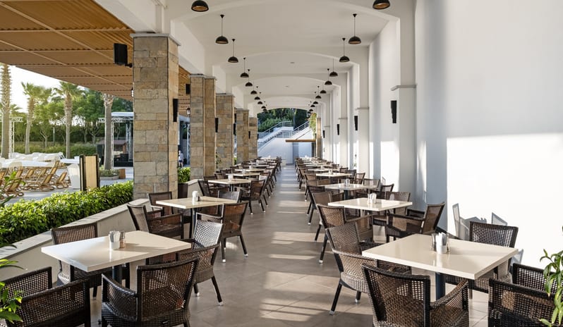 Xanadu Bar An elegant outdoor dining terrace at a seaside resort featuring stone pillars, wicker seating, and a view of a pool and mountains by the ocean at Xanadu Island by Kenwood Travel