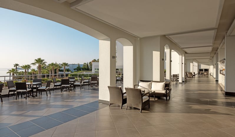 Rosebud Bar & Terrace Spacious shaded outdoor terrace of a luxury resort featuring wicker lounge seating, white arched pillars, and a clear view of palm trees by the ocean at Xanadu Island by Kenwood Travel