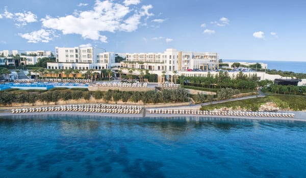 Beach Bar Aerial view of a luxury white-domed resort in Crete featuring a long turquoise swimming pool, rows of beach lounge chairs, and a private sandy cove at Xanadu Island by Kenwood Travel