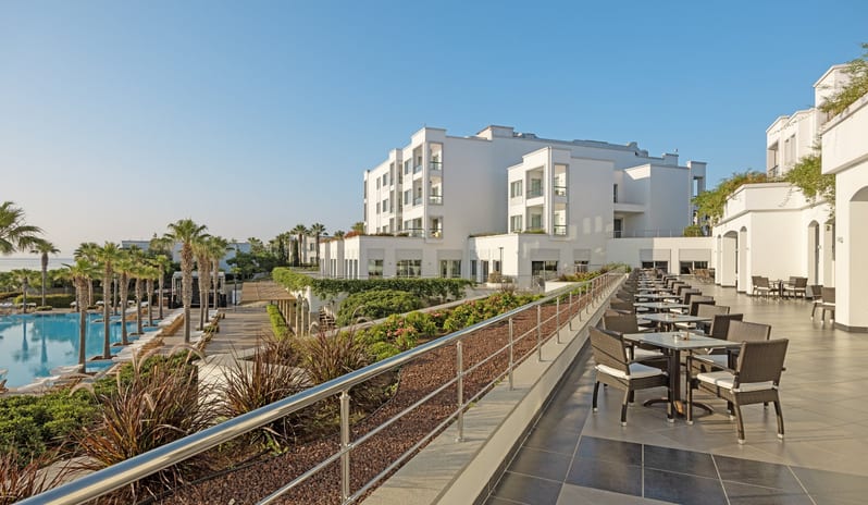 Rosebud Bar & Terrace Spacious outdoor terrace of a white luxury beach resort featuring cafe seating, a large swimming pool lined with palm trees, and clear blue skies at Xanadu Island by Kenwood Travel