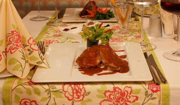 Azzurro à la Carte Restaurant close-up of a gourmet roasted meat dish covered in brown gravy on a white rectangular plate, set on a floral patterned table runner with wine glasses and breadsticks in the background at Xanadu Island by Kenwood Travel