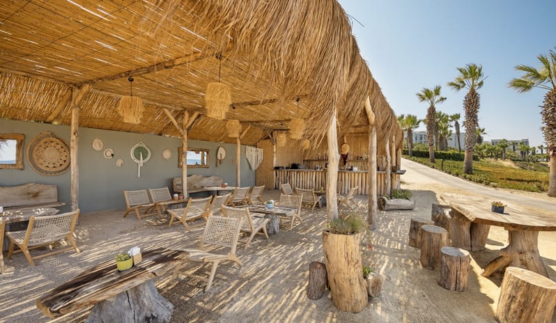 Abora Beach Bar A rustic outdoor beach bar with a thatched roof, wooden log seating, and bohemian decor under a clear blue sky at Xanadu Island by Kenwood Travel