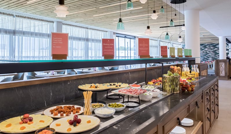 Roots Buffet Restaurant featuring a variety of sliced cheeses, cured meats, fresh fruits, and olives arranged on a dark marble counter under warm pendant lighting at Barcelo Nura by Kenwood Travel