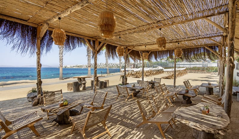 Abora Beach Bar A rustic beach club lounge with a thatched bamboo roof, live-edge wooden tables, and woven chairs on a sandy beach overlooking the oceanat Xanadu Island by Kenwood Travel