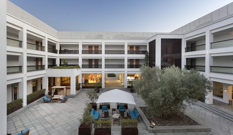 Lobby Bar A modern white three-story luxury resort courtyard with teal outdoor seating, a central olive tree, and boutique shops under a clear blue sky at Xanadu Island by Kenwood Travel