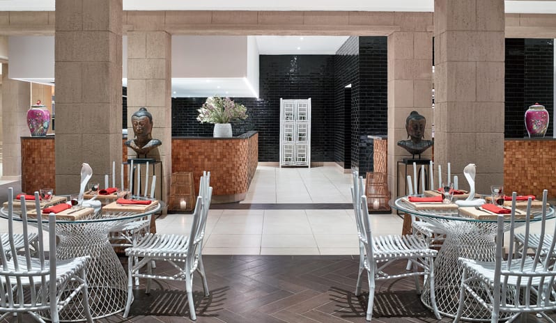 Asiana white wire chairs, glass tables with red napkins, and Asian-inspired decor including Buddha busts and ceramic vases at Grecotel LUXME Dama Dama by Kenwood Travel