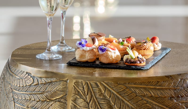 The RoseTwo glasses of chilled champagne and a selection of gourmet pastries on a decorative bronze table at Grecotel LUXME Dama Dama by Kenwood Travel