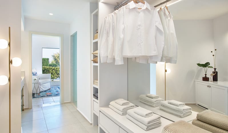 LUX ME Family Bungalow Pool side organized shelving, folded towels, and hanging white shirts with a view into the living area at Grecotel LUXME Dama Dama by Kenwood Travel