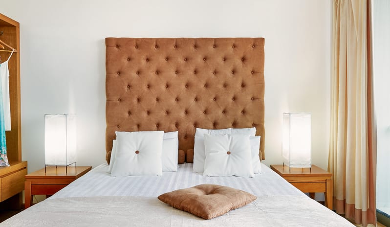 PLUX ME Family Guestroom Sea View bright hotel bedroom with a large brown tufted headboard, white pillows, and two glowing bedside lamp at Grecotel LUXME Dama Dama by Kenwood Travel