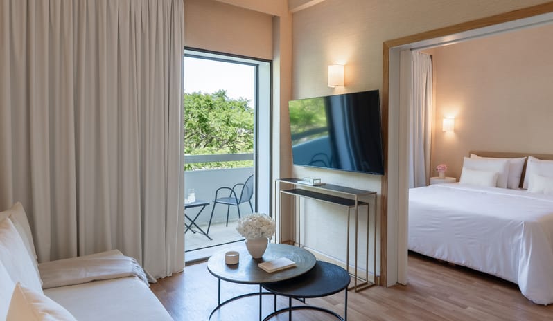 Superior Family Apartment Inland View modern living room with a wall-mounted TV, a separate bedroom with white linens, and a balcony overlooking green trees at Grecotel LUXME Dama Dama by Kenwood Travel