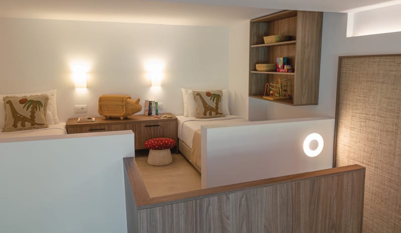Premier Loft Family Guestroom twin beds with dinosaur pillows, a whimsical mushroom stool, and curated toys on wooden shelving at Grecotel LUXME Dama Dama by Kenwood Travel