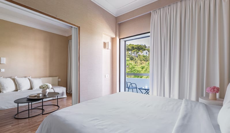 Superior Family Apartment Inland View primary bedroom with white linens, a separate living area with a sofa, and a private balcony overlooking inland greenery at Grecotel LUXME Dama Dama by Kenwood Travel