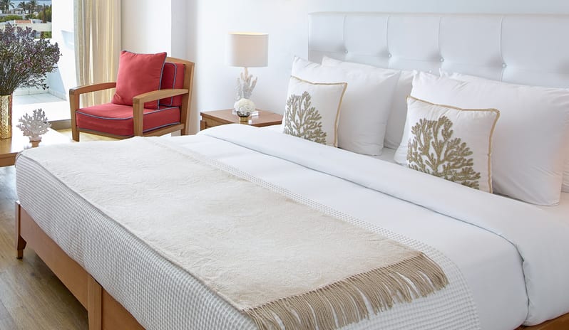 Superior Guestroom Side Sea View white bedding with coral-patterned pillows and a red accent chair at Grecotel LUXME Dama Dama by Kenwood Travel
