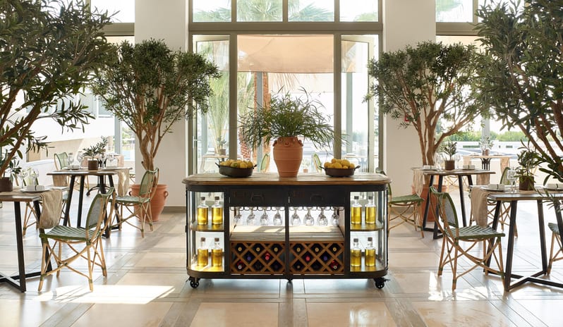Italian à la Carte Sunlit Mediterranean restaurant interior with olive trees, lemon displays, and a vintage-style wine and spirits cart at Grecotel LUXME Dama Dama by Kenwood Travel