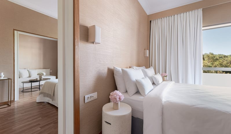 Superior Family Apartment Inland View primary bedroom with white linens, a separate living area with a sofa, and a private balcony overlooking inland greenery at Grecotel LUXME Dama Dama by Kenwood Travel