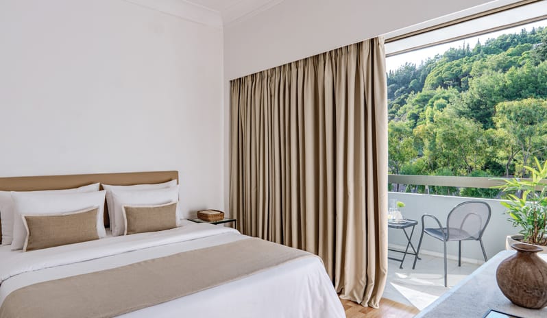 Premier Family Guestroom Inland View white linens, neutral accents, and a private balcony with a view of lush green trees at Grecotel LUXME Dama Dama by Kenwood Travel