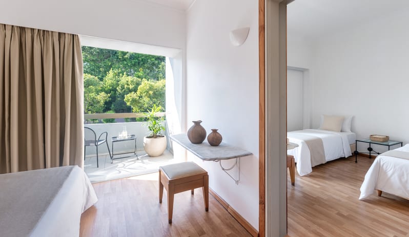 Premier Family Guestroom Inland View twin twin beds, minimalist white decor, and a view into the main room with a private balconyat Grecotel LUXME Dama Dama by Kenwood Travel