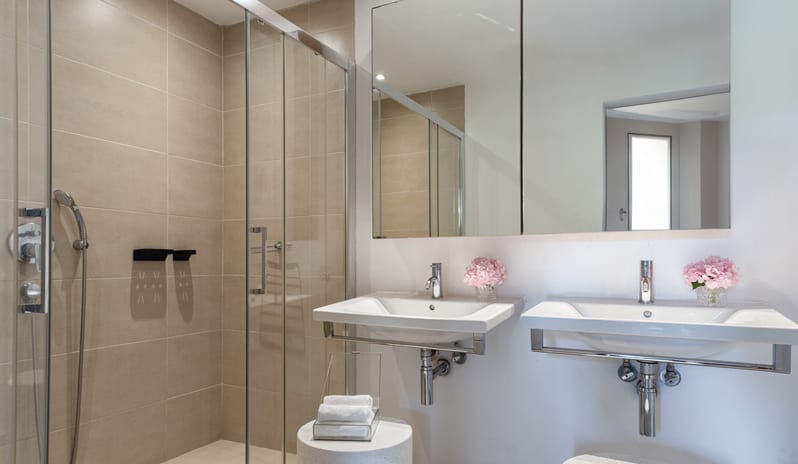 Superior Family Apartment Inland View walk-in glass shower, double sinks with chrome fixtures, large mirrors, and decorative pink flowers at Grecotel LUXME Dama Dama by Kenwood Travel