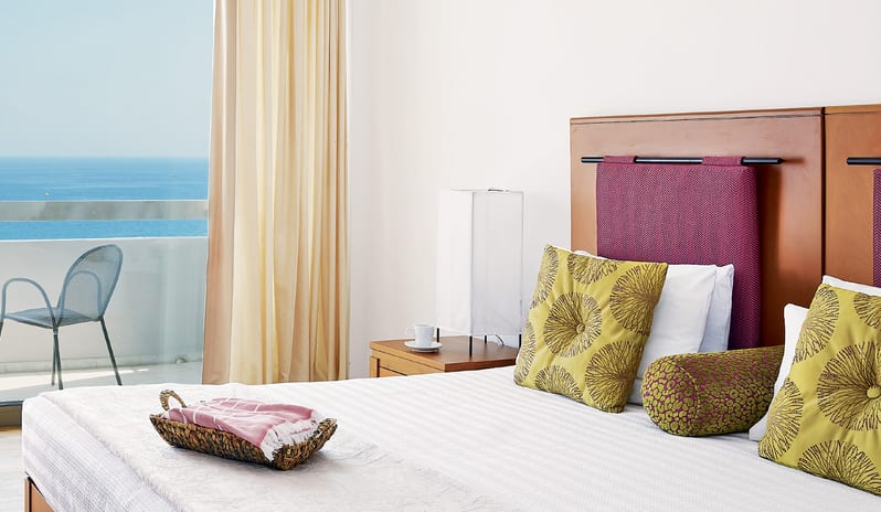 Superior Guestroom Sea View private balcony with a sea view, colorful patterned pillows, and a white striped bedspread at Grecotel LUXME Dama Dama by Kenwood Travel
