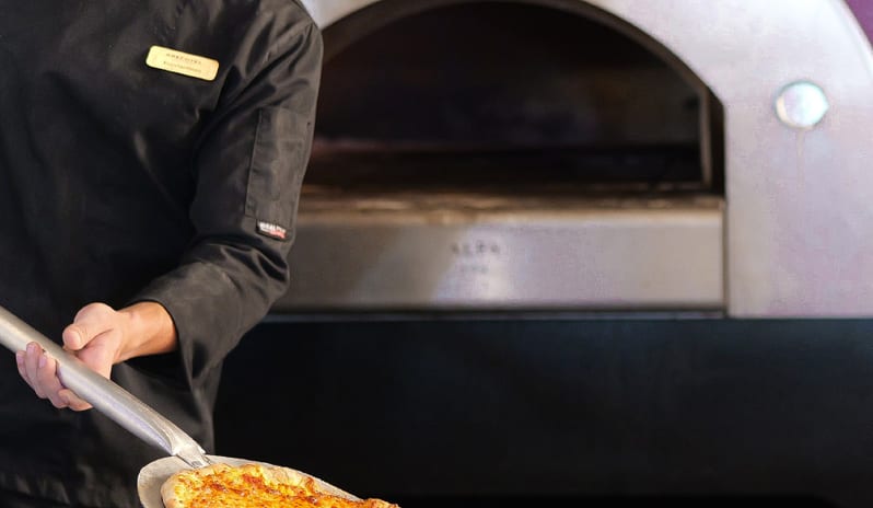 Dama Dama A chef in a black uniform using a pizza peel to remove a fresh, golden-brown cheese pizza from a traditional wood-fired oven at Grecotel LUXME Dama Dama by Kenwood Travel