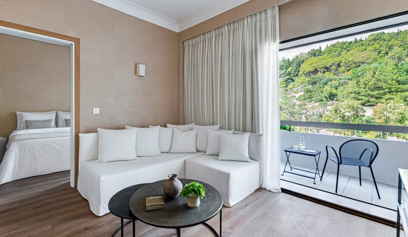 Superior Family Apartment Inland View modern living area with a white sectional sofa, a glimpse of the bedroom, and a private balcony overlooking a lush green hillside at Grecotel LUXME Dama Dama by Kenwood Travel