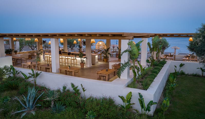 Beach Club wooden tables, woven pendant lights, and a view of the Aegean Sea at dusk at Grecotel LUXME Dama Dama by Kenwood Travel